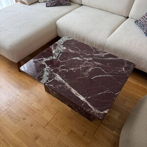 May include: A square coffee table with a dark burgundy marble top and a matching base. The marble has white veining. The table is in a living room setting with a cream-colored sectional sofa and hardwood flooring.