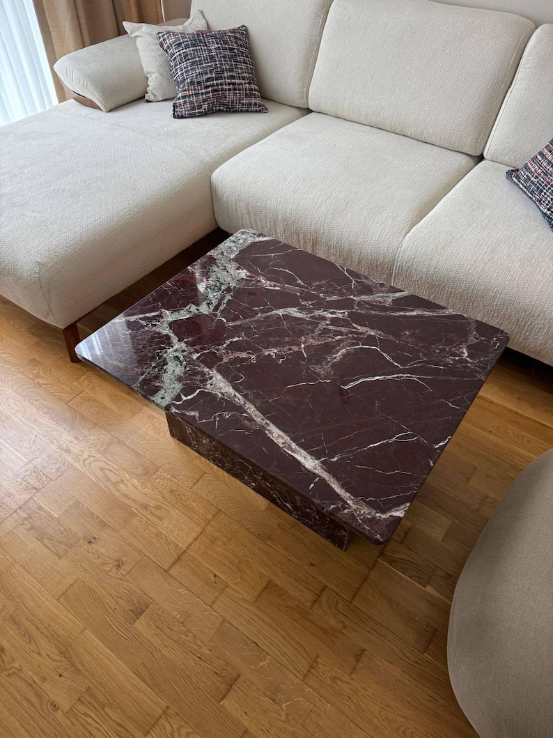 May include: A square coffee table with a dark burgundy marble top and a matching base. The marble has white veining. The table is in a living room setting with a cream-colored sectional sofa and hardwood flooring.