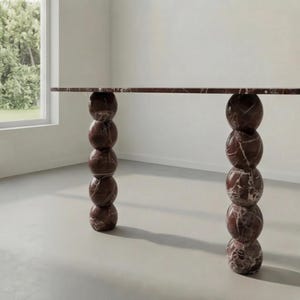 May include: A modern, rectangular table with a reddish-brown marble top. The table is supported by two sets of stacked, spherical legs. Each leg is composed of five marble spheres. The table is in a room with a window.