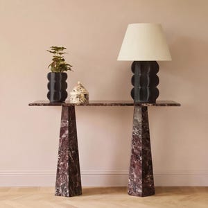 Könnte beinhalten: Eine moderne Konsole mit rechteckiger Marmorplatte und zwei trapezförmigen Beinen. Eine schwarze Lampe mit cremefarbenem Lampenschirm, eine schwarze Vase und ein dekoratives Glas stehen auf dem Tisch. Tisch und Beine sind tiefrot mit weißen Adern.