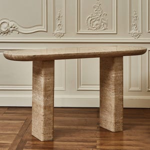 Puede incluir: Una consola de piedra beige con una parte superior rectangular redondeada y dos patas rectangulares. La mesa está hecha de piedra de color claro y tiene un acabado liso. La mesa mide aproximadamente 152 cm de largo.