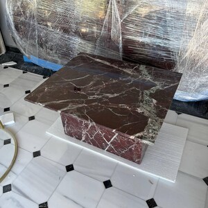 May include: A square, dark red marble coffee table with white veining. The table is supported by a rectangular base of the same material. The table is sitting on a white surface, with a marble tile floor in the background.