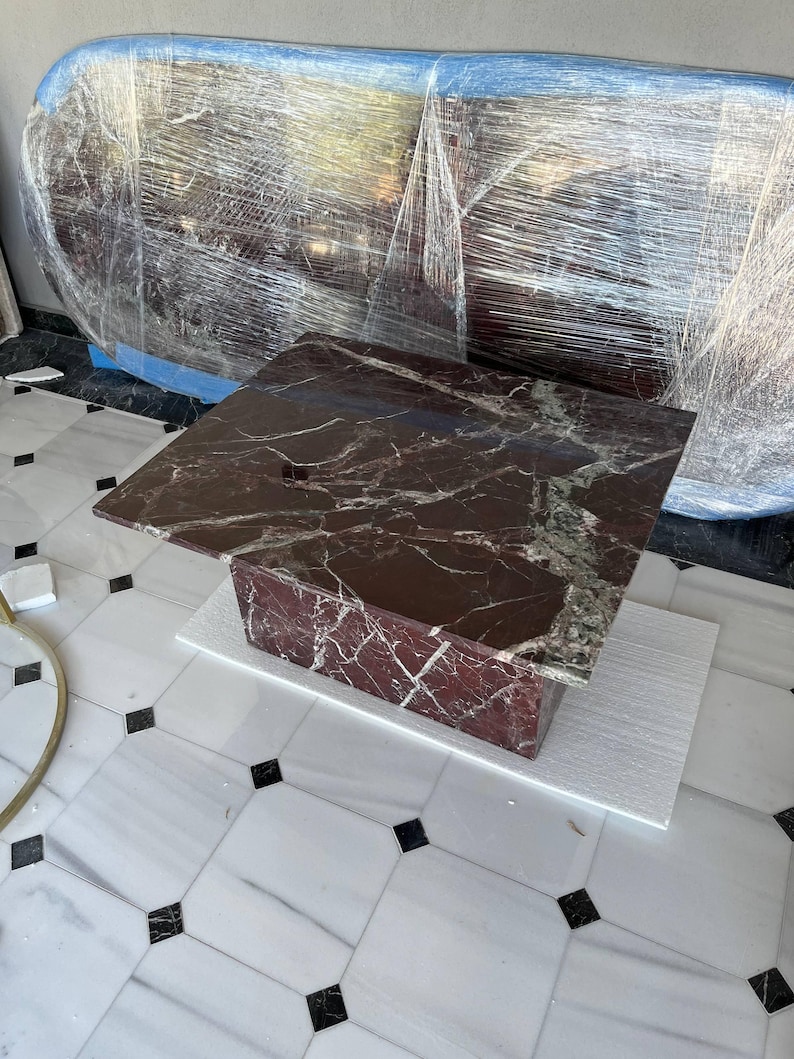 May include: A square, dark red marble coffee table with white veining. The table is supported by a rectangular base of the same material. The table is sitting on a white surface, with a marble tile floor in the background.