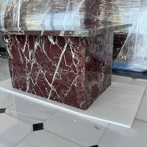 May include: A rectangular marble table with a deep red and white veined pattern. The table is wrapped in clear plastic. The table is sitting on a white surface, and the floor is white tile with black and gray accents.