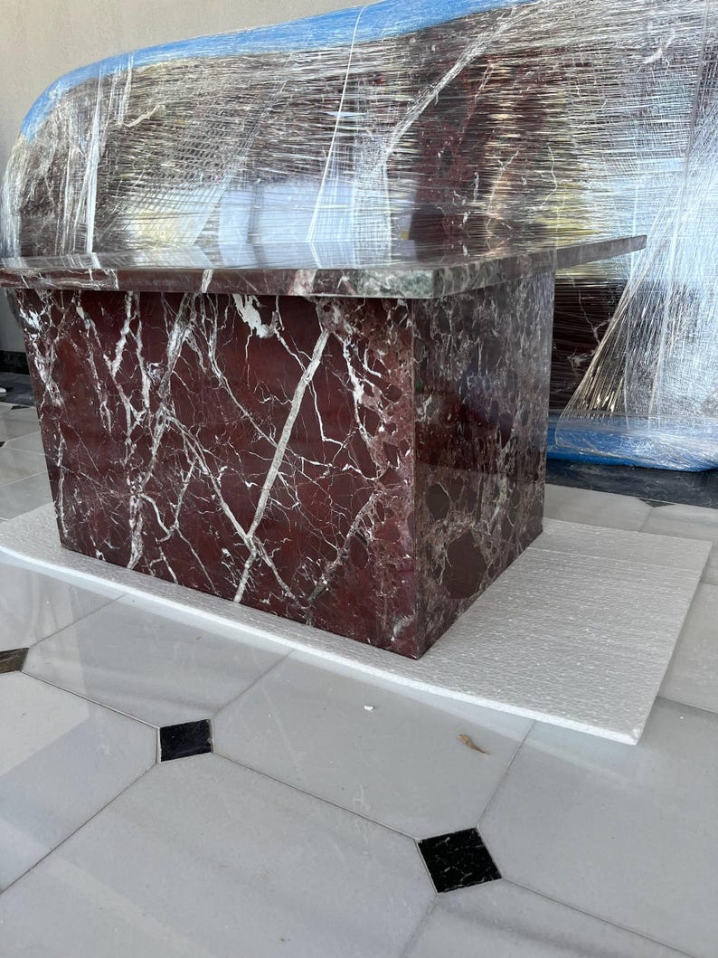 May include: A rectangular marble table with a deep red and white veined pattern. The table is wrapped in clear plastic. The table is sitting on a white surface, and the floor is white tile with black and gray accents.