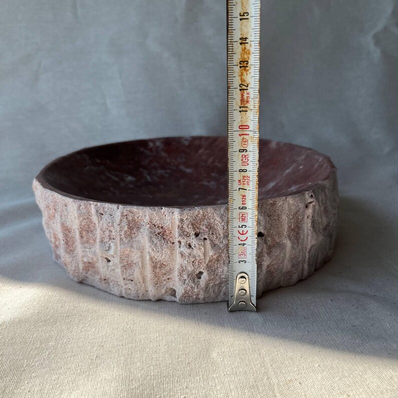 Puede incluir: Un cuenco de piedra tallado a mano con un exterior rugoso y texturizado y un interior liso y rojo oscuro. El cuenco tiene aproximadamente 12 cm de di&aacute;metro y 7,5 cm de alto. El exterior es de color beige claro.