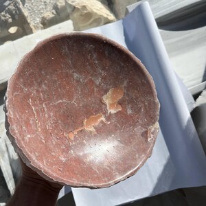 Puede incluir: Un cuenco redondo de piedra de color marr&oacute;n rojizo con una superficie rugosa y texturizada. El cuenco tiene poca profundidad y un borde ligeramente irregular. El interior presenta marcas m&aacute;s claras y unas peque&ntilde;as manchas irregulares de un tono m&aacute;s claro.