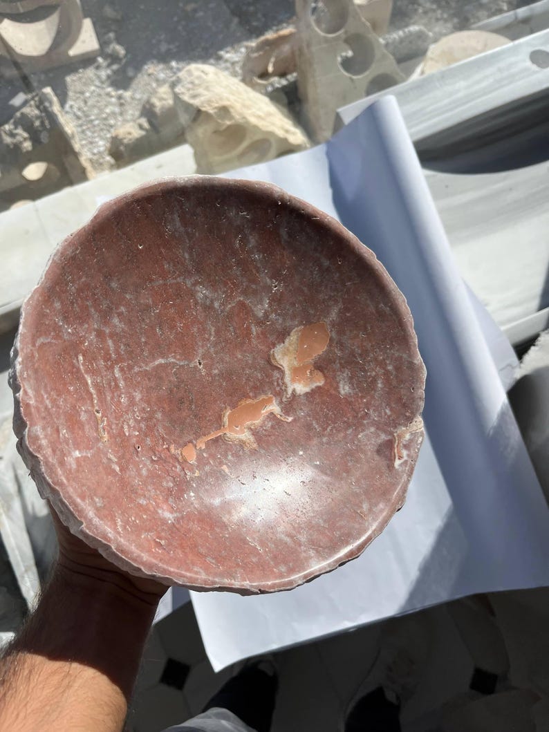 Puede incluir: Un cuenco redondo de piedra de color marr&oacute;n rojizo con una superficie rugosa y texturizada. El cuenco tiene poca profundidad y un borde ligeramente irregular. El interior presenta marcas m&aacute;s claras y unas peque&ntilde;as manchas irregulares de un tono m&aacute;s claro.