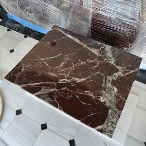 May include: A square, dark red marble tabletop with white and green veining. The marble is on a white surface with black square accents. The tabletop is wrapped in clear plastic.