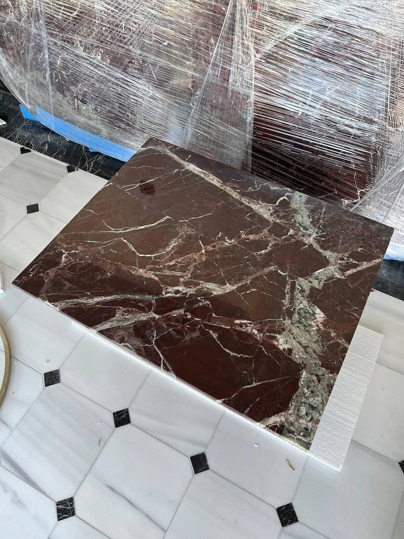 May include: A square, dark red marble tabletop with white and green veining. The marble is on a white surface with black square accents. The tabletop is wrapped in clear plastic.