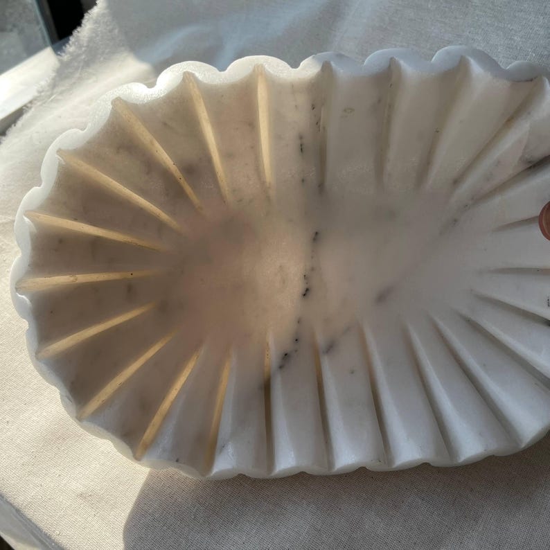 May include: A white marble decorative bowl with a scalloped edge and a fluted design. The oval-shaped bowl has subtle gray veining and is set against a neutral background. This elegant piece is suitable for home decor.
