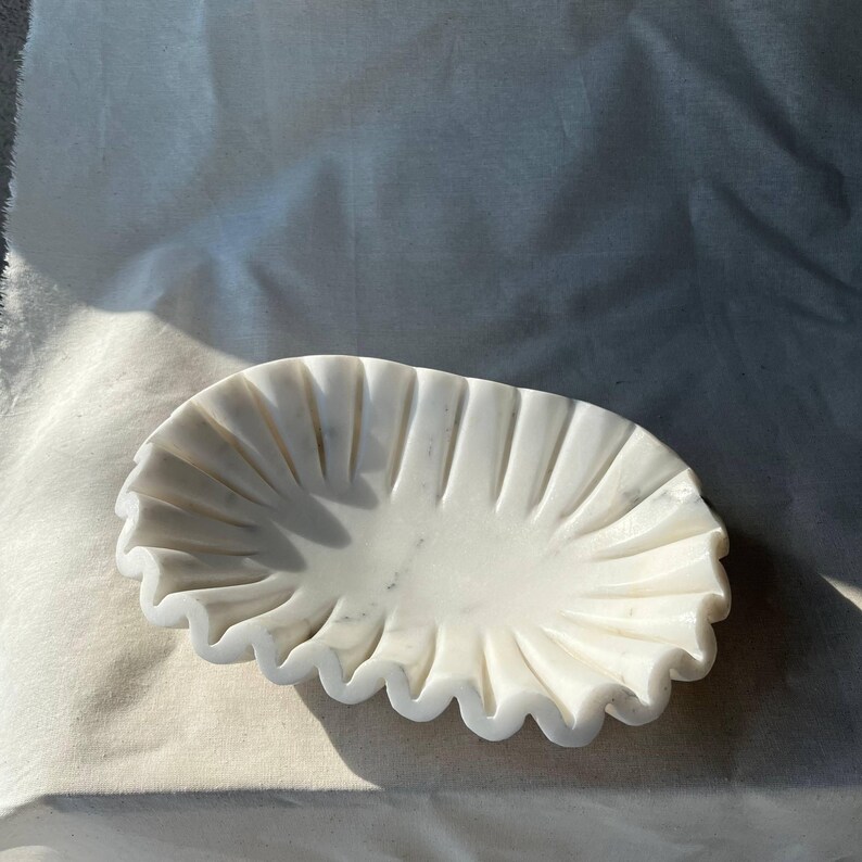 May include: A white marble decorative bowl with a ruffled edge. The bowl has a shallow, oval shape and is carved with vertical lines radiating from the center. The marble has subtle gray veining, adding to its natural beauty.