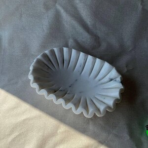 May include: A white marble decorative dish with a scalloped edge. The oval-shaped dish has a fluted design radiating from the center. The marble has subtle gray veining. The dish is on a neutral-colored surface.
