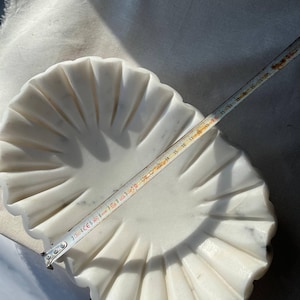 May include: A white marble decorative bowl with a scalloped edge design. The bowl is approximately 10 inches in diameter. The bowl has a fluted pattern radiating from the center.