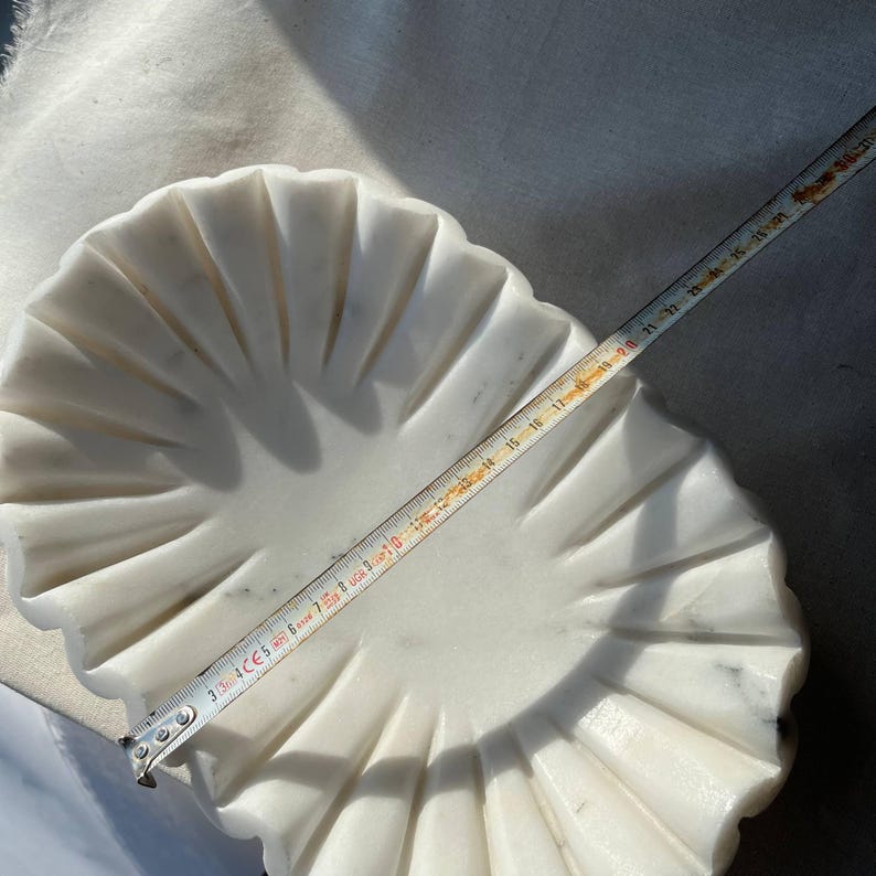 May include: A white marble decorative bowl with a scalloped edge design. The bowl is approximately 10 inches in diameter. The bowl has a fluted pattern radiating from the center.