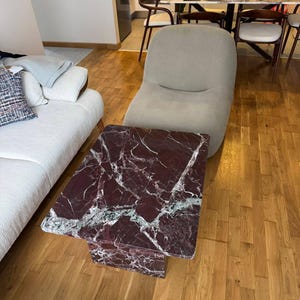 May include: A rectangular coffee table with a burgundy marble top and a single pedestal base. The marble has white and green veining. The table is in a living room with a beige chair and hardwood floors.