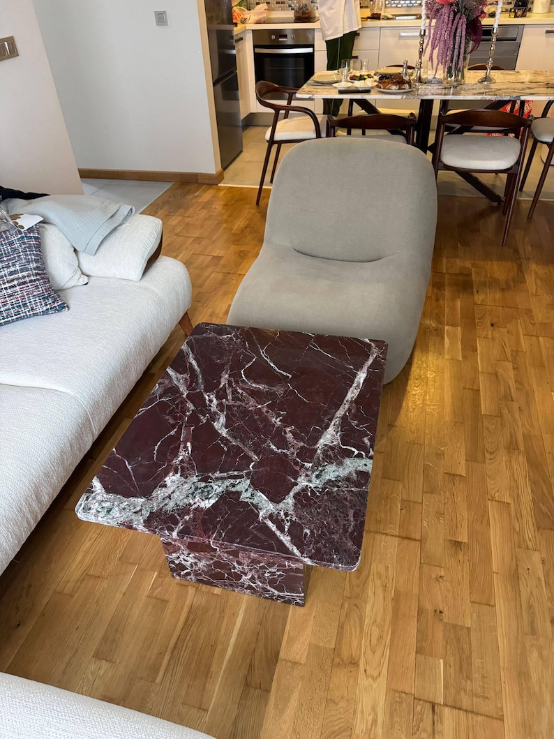 May include: A rectangular coffee table with a burgundy marble top and a single pedestal base. The marble has white and green veining. The table is in a living room with a beige chair and hardwood floors.