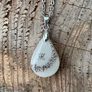 May include: A silver chain necklace with a teardrop-shaped pendant. The pendant is white with a glitter border and a black letter "M" in the center. The necklace is displayed on a wooden surface.