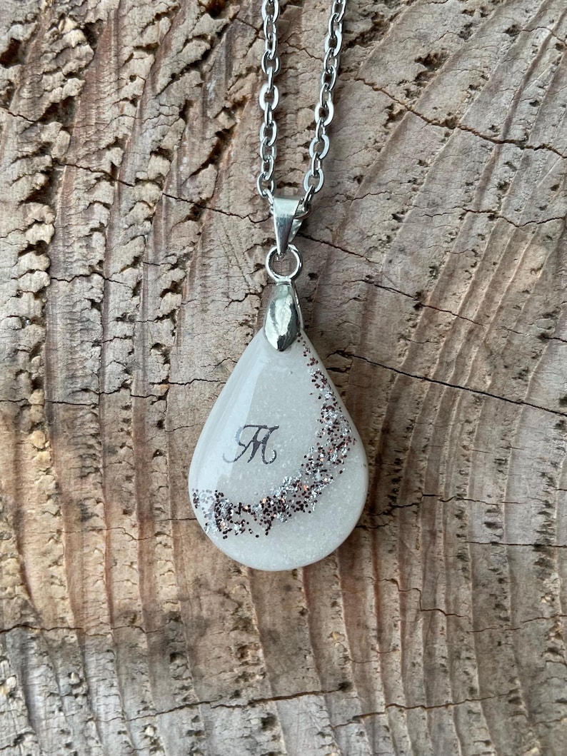 May include: A silver chain necklace with a teardrop-shaped pendant. The pendant is white with a glitter border and a black letter "M" in the center. The necklace is displayed on a wooden surface.