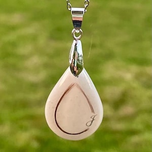 May include: A silver necklace with a teardrop-shaped pendant. The pendant is a light pink color with a silver outline and a cursive letter "J" engraved on the front. The background is blurred green grass.