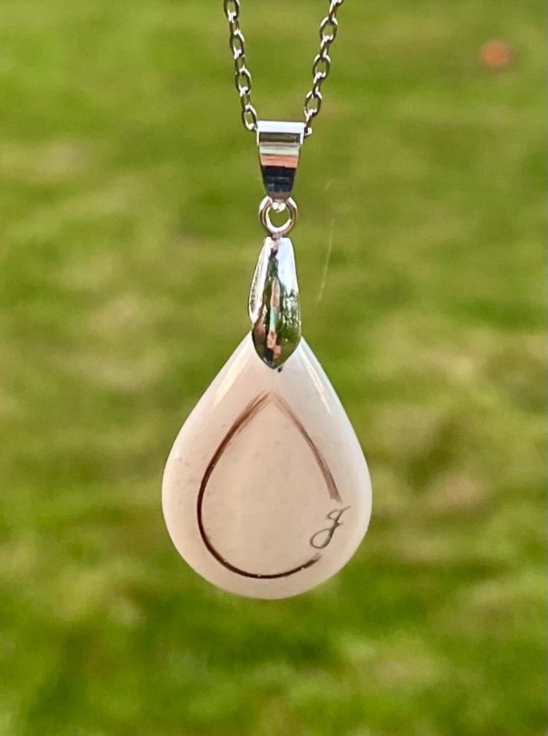 May include: A silver necklace with a teardrop-shaped pendant. The pendant is a light pink color with a silver outline and a cursive letter "J" engraved on the front. The background is blurred green grass.