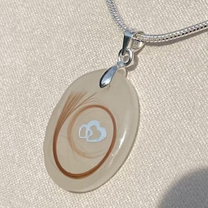 May include: A silver necklace with a pendant. The pendant is an oval shape with a clear, light-colored resin. Inside the resin are strands of brown hair and a light blue double heart design. The necklace chain is silver.