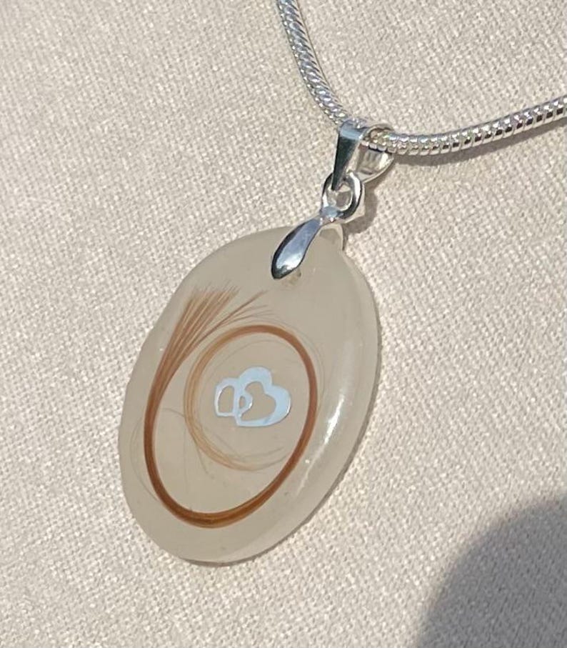 May include: A silver necklace with a pendant. The pendant is an oval shape with a clear, light-colored resin. Inside the resin are strands of brown hair and a light blue double heart design. The necklace chain is silver.
