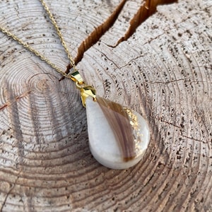 May include: A gold-colored chain necklace with a teardrop pendant. The pendant is white with a section of brown hair and gold leaf accents. The necklace is displayed on a textured wooden surface.