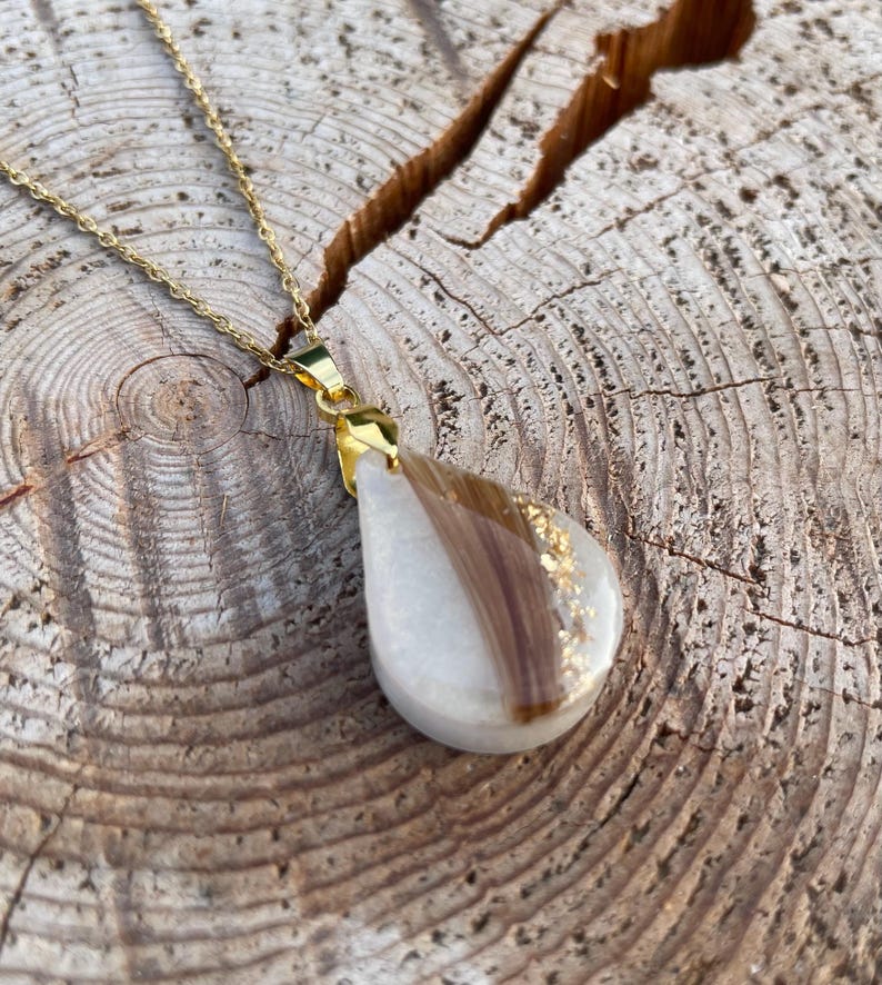 May include: A gold-colored chain necklace with a teardrop pendant. The pendant is white with a section of brown hair and gold leaf accents. The necklace is displayed on a textured wooden surface.