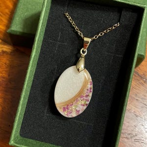 May include: A gold-colored chain necklace with an oval pendant. The pendant features a white background with a curved section of light brown material and pink floral accents. The necklace is presented in a green box with a black interior.