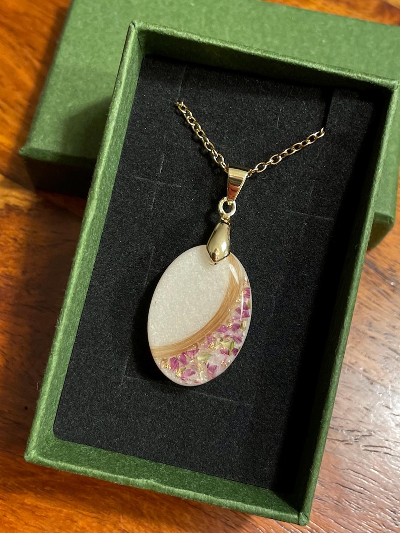 May include: A gold-colored chain necklace with an oval pendant. The pendant features a white background with a curved section of light brown material and pink floral accents. The necklace is presented in a green box with a black interior.