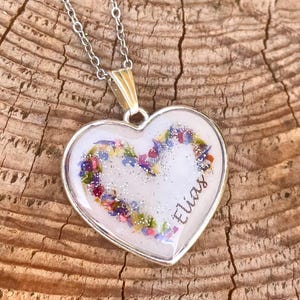Può includere: Collana con pendente a forma di cuore argentato. Il pendente presenta uno sfondo bianco con fiori secchi colorati disposti a forma di cuore. Il nome "Elias" è scritto in corsivo sul pendente.