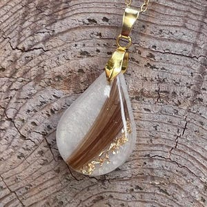May include: A teardrop-shaped pendant necklace with a gold chain. The pendant is clear with a section of brown hair and gold flakes. The pendant is attached to a gold bail.