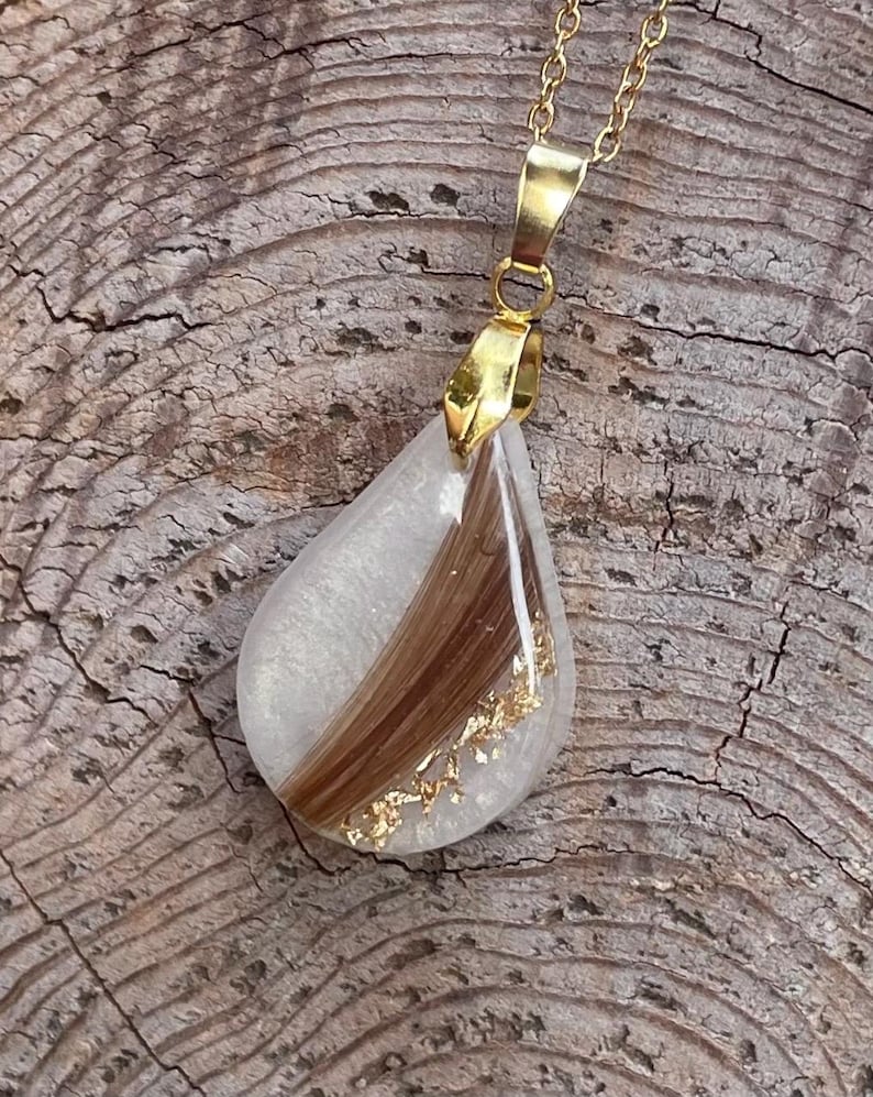 May include: A teardrop-shaped pendant necklace with a gold chain. The pendant is clear with a section of brown hair and gold flakes. The pendant is attached to a gold bail.