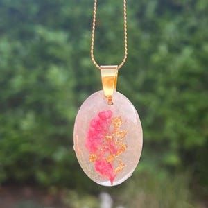 May include: Oval pendant necklace with a gold chain. The pendant is clear with a pink floral design and gold flakes. The pendant is suspended from a gold-colored bail.