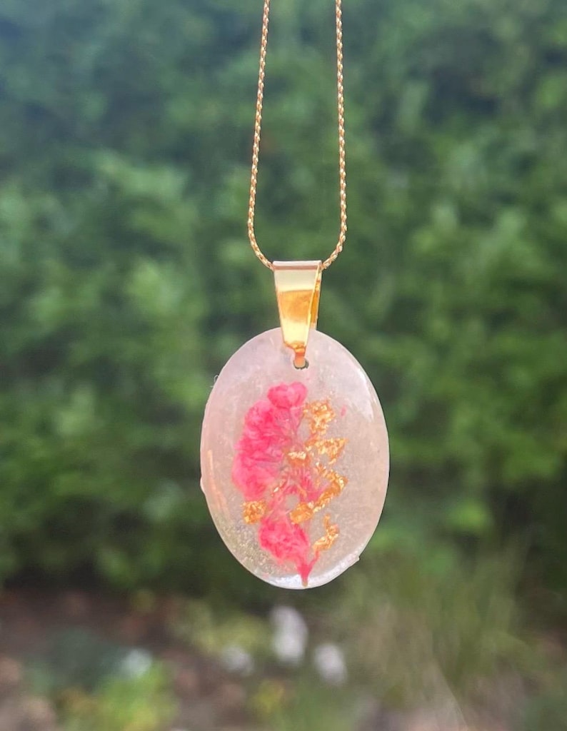 May include: Oval pendant necklace with a gold chain. The pendant is clear with a pink floral design and gold flakes. The pendant is suspended from a gold-colored bail.