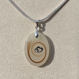May include: Oval pendant necklace with a silver chain. The pendant is a cream color with a swirl of brown hair and two black hearts. The necklace is a delicate piece of jewelry.