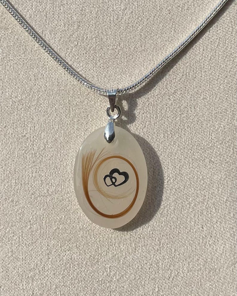 May include: Oval pendant necklace with a silver chain. The pendant is a cream color with a swirl of brown hair and two black hearts. The necklace is a delicate piece of jewelry.