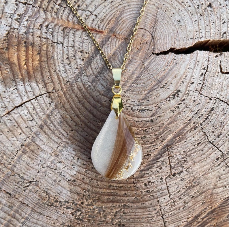 May include: A gold-colored chain necklace with a teardrop pendant. The pendant is white with a section of brown hair and gold leaf accents. The necklace is displayed on a wooden surface.