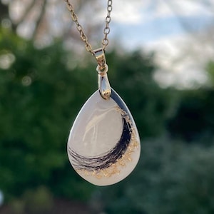 May include: A teardrop-shaped pendant necklace with a gold chain. The pendant features a white background with black and gold accents. The necklace is a delicate piece of jewelry.