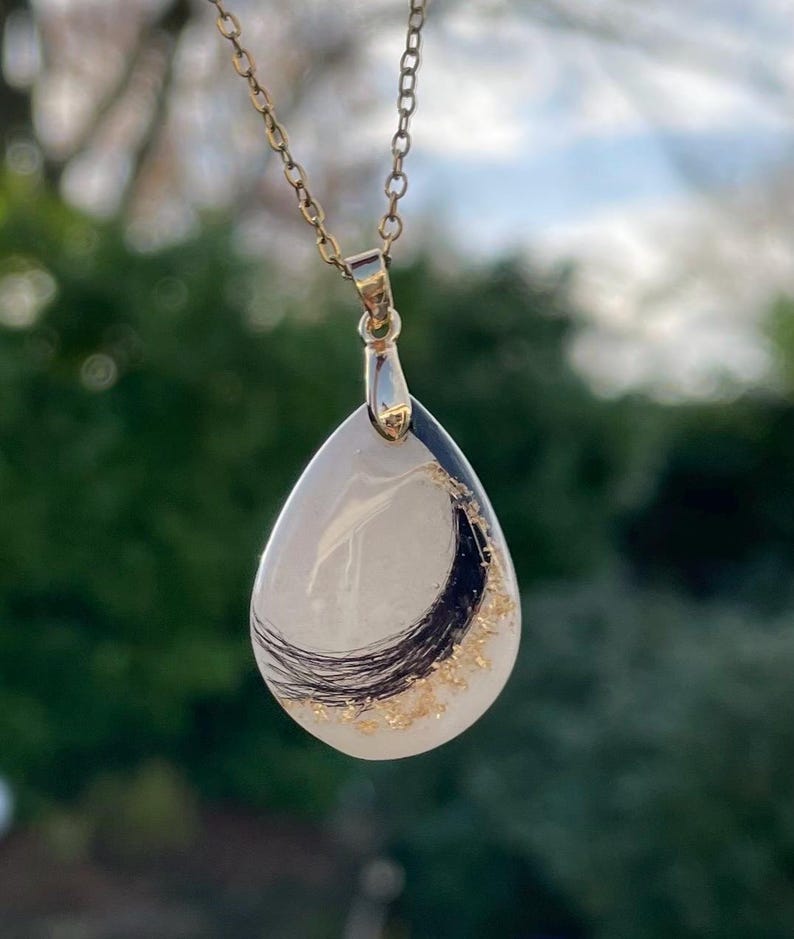 May include: A teardrop-shaped pendant necklace with a gold chain. The pendant features a white background with black and gold accents. The necklace is a delicate piece of jewelry.