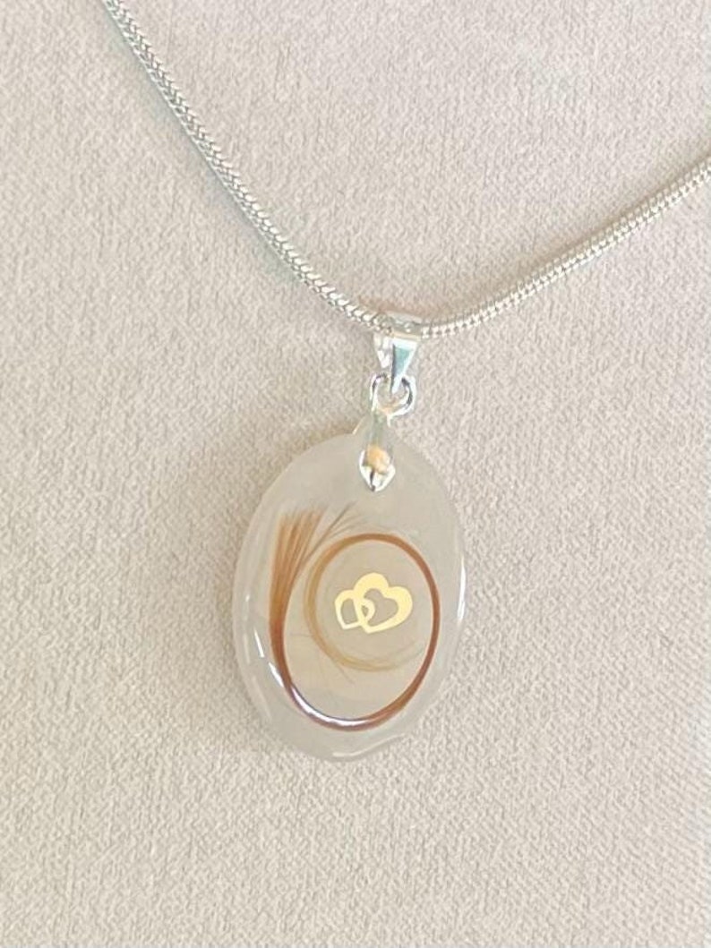 May include: A silver necklace with a pendant featuring a light-colored oval resin. Inside the resin is a swirl of brown hair and a gold-colored double heart design. The necklace chain is a thin, silver snake chain.