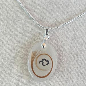 May include: A silver necklace with a pendant. The pendant is an oval shape with a clear resin. Inside the resin is a swirl of brown hair and a black heart design. The chain is a silver snake chain.