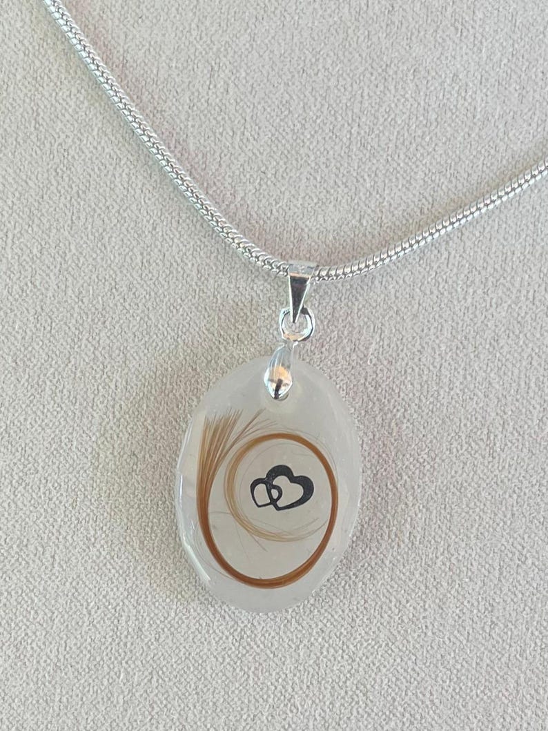 May include: A silver necklace with a pendant. The pendant is an oval shape with a clear resin. Inside the resin is a swirl of brown hair and a black heart design. The chain is a silver snake chain.