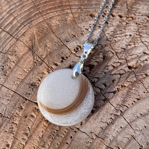 May include: A round pendant necklace with a silver chain. The pendant features a white background with a curved section of brown hair and silver glitter. The pendant is attached to a silver bail.