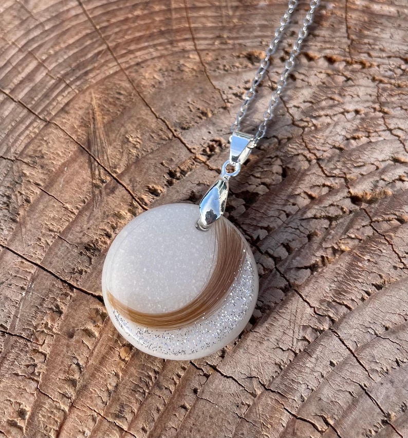 May include: A round pendant necklace with a silver chain. The pendant features a white background with a curved section of brown hair and silver glitter. The pendant is attached to a silver bail.