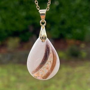 May include: A gold-colored chain necklace with a teardrop pendant. The pendant is clear with strands of brown and auburn hair and gold flakes. The background is blurred green.