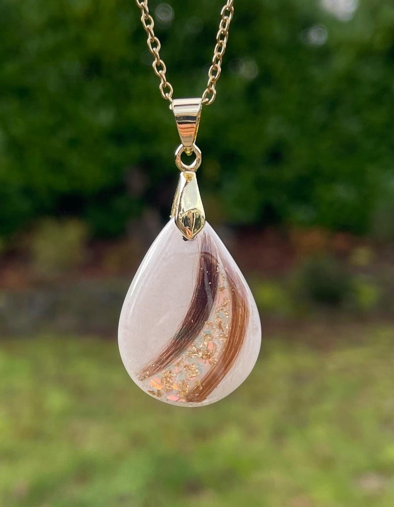 May include: A gold-colored chain necklace with a teardrop pendant. The pendant is clear with strands of brown and auburn hair and gold flakes. The background is blurred green.