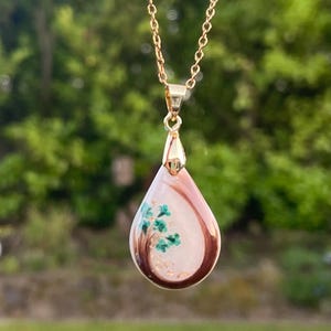 May include: Gold-toned necklace with a teardrop pendant. The pendant features a light pink and white background with a brown border. Inside the pendant is a small green tree design with gold flecks. The necklace is displayed against a blurred green background.