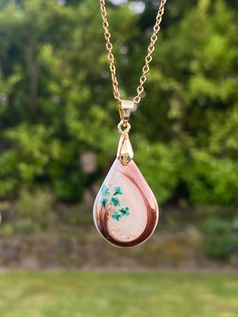 May include: Gold-toned necklace with a teardrop pendant. The pendant features a light pink and white background with a brown border. Inside the pendant is a small green tree design with gold flecks. The necklace is displayed against a blurred green background.